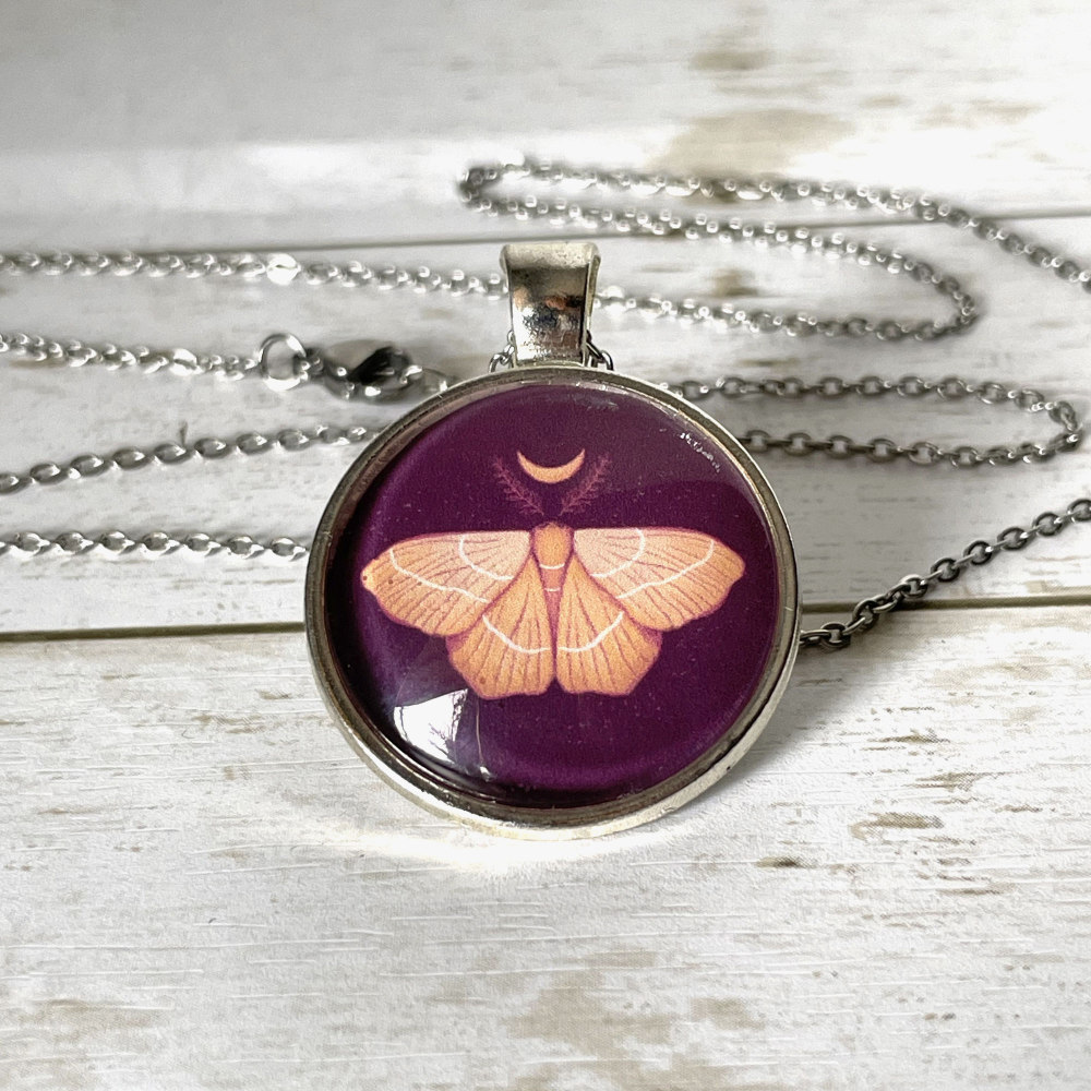 Handmade cabochon necklace with stainless steel chain and original art of an orange moth and moon against dark purple background.