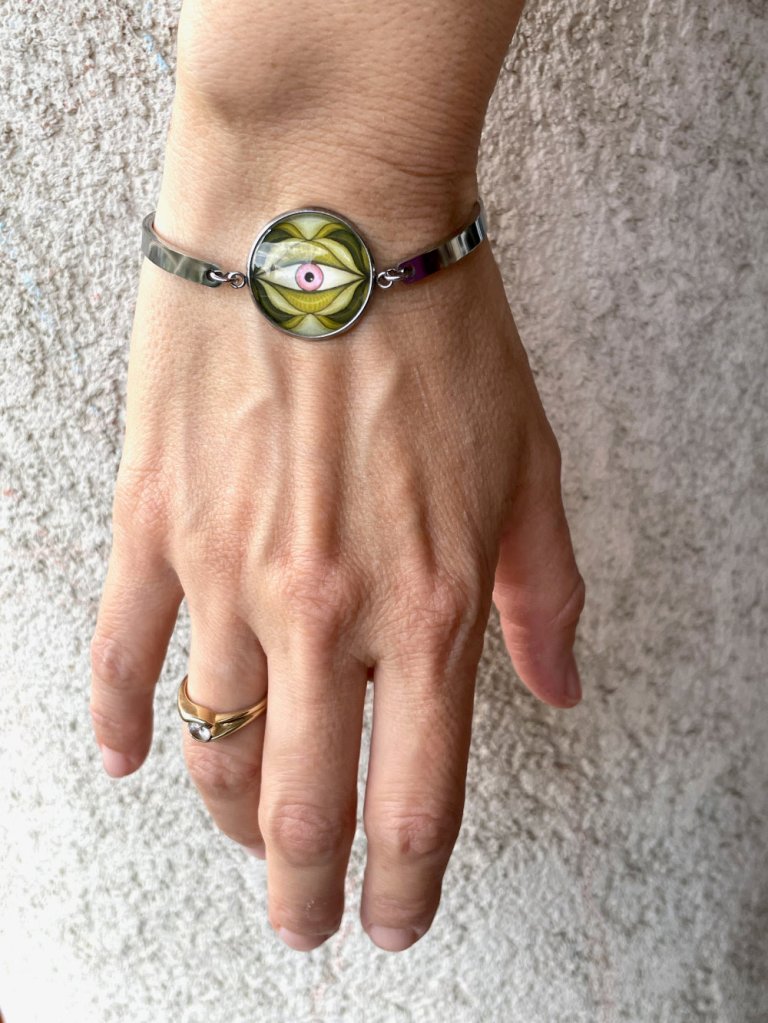 Гривна със странно око в зелено и розово за дами с нестандартен вкус. Изработена от неръждаема стомана и стъкло, с регулируема верижка.