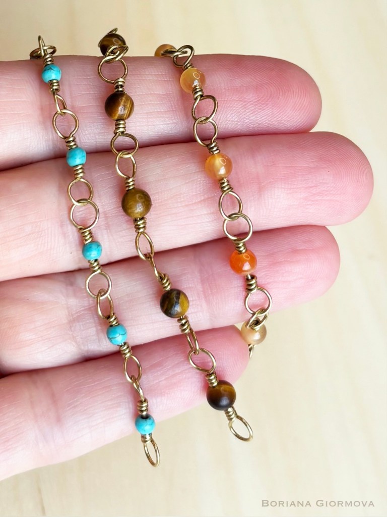 Three unfinished handmade brass chains held in hand - one with carnelian, one with tiger’s eye and one with turquoise stones