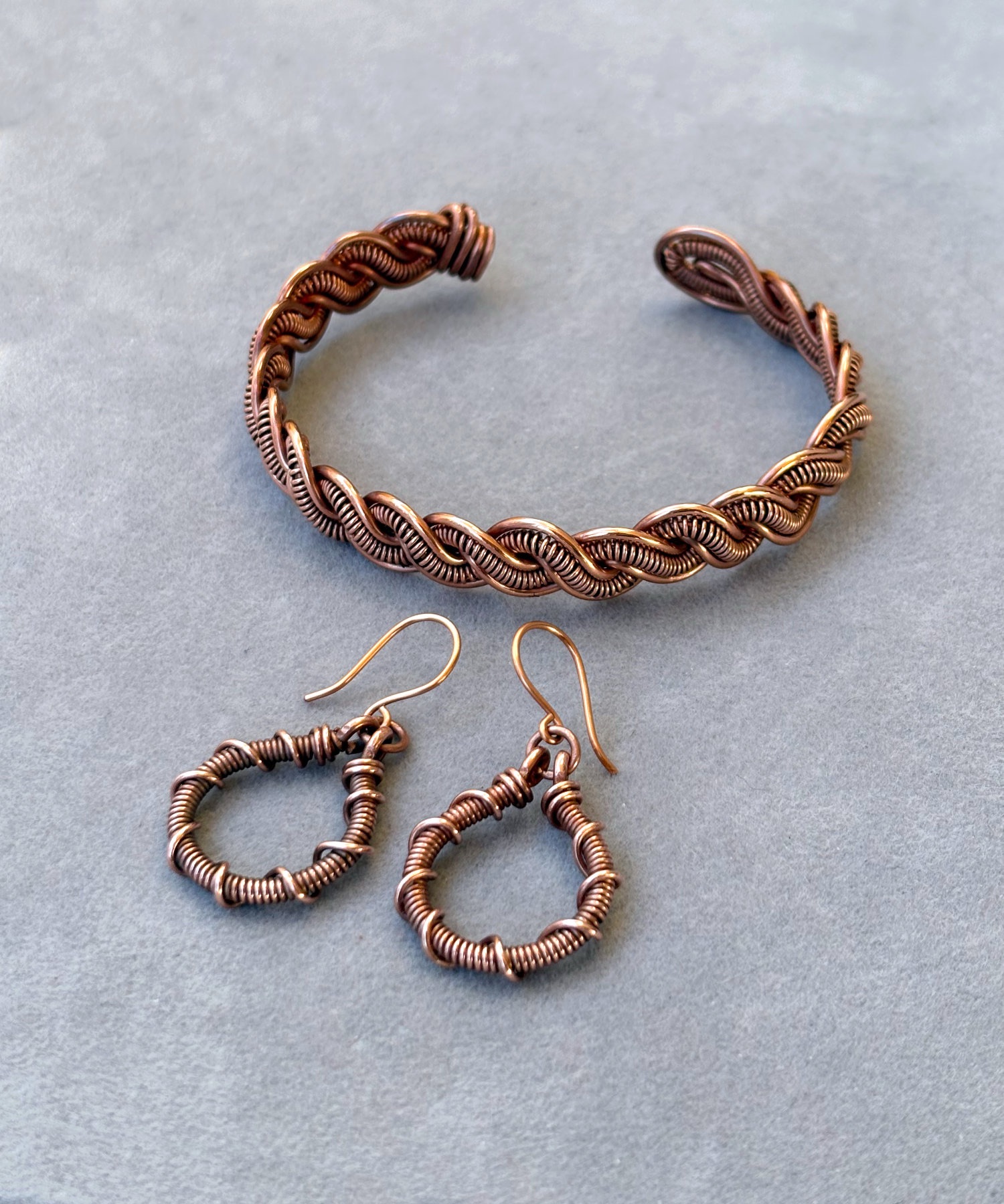 A matching set of handmade jewelry including a wide woven copper cuff bracelet and copper wire-wrapped drop earrings on a neutral background.