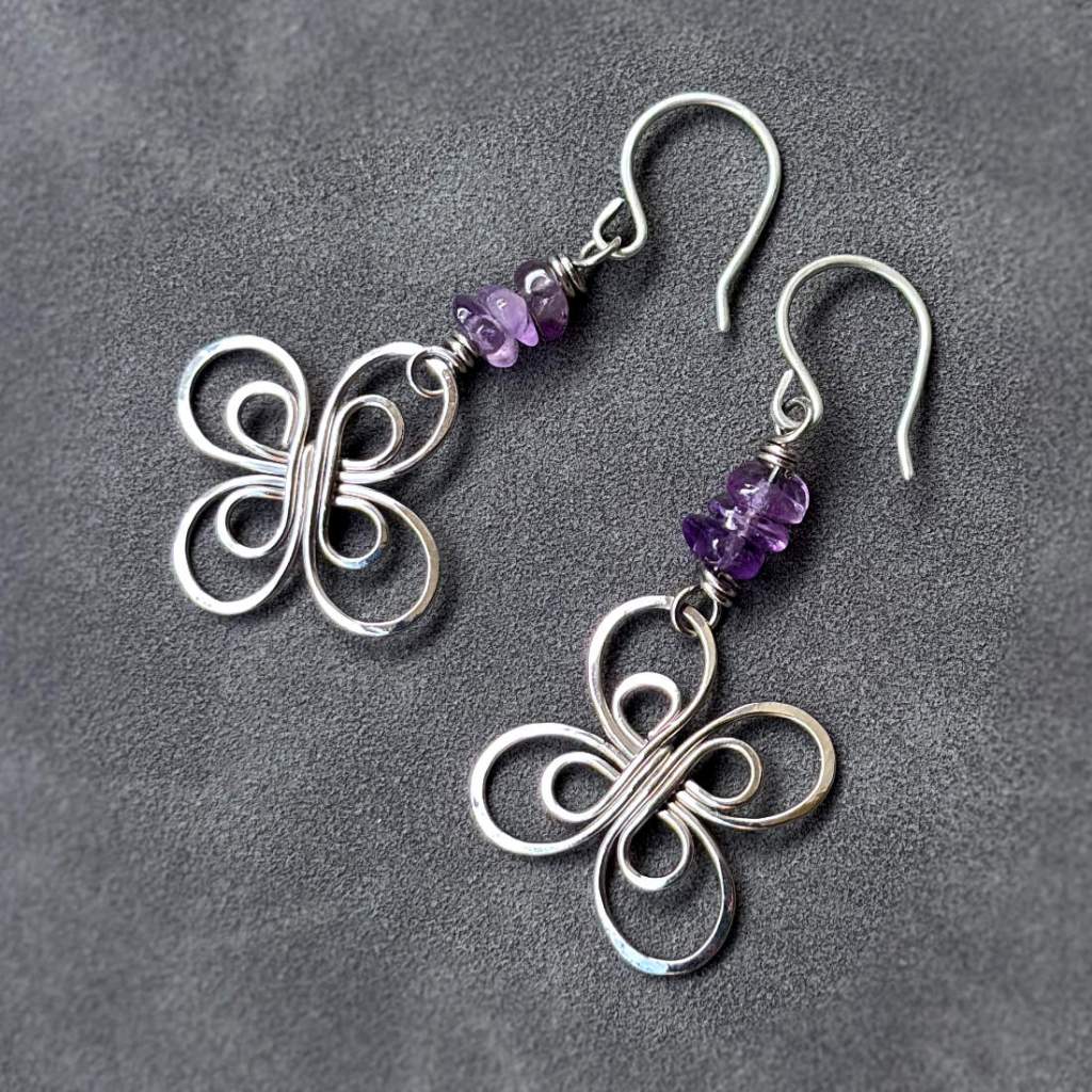 Сребристи обици с форма на елеганнтно цвете, свързано с три камъка лилав аметист чипс, висящи от сребърни кукички, снимани на тъмносив велур.