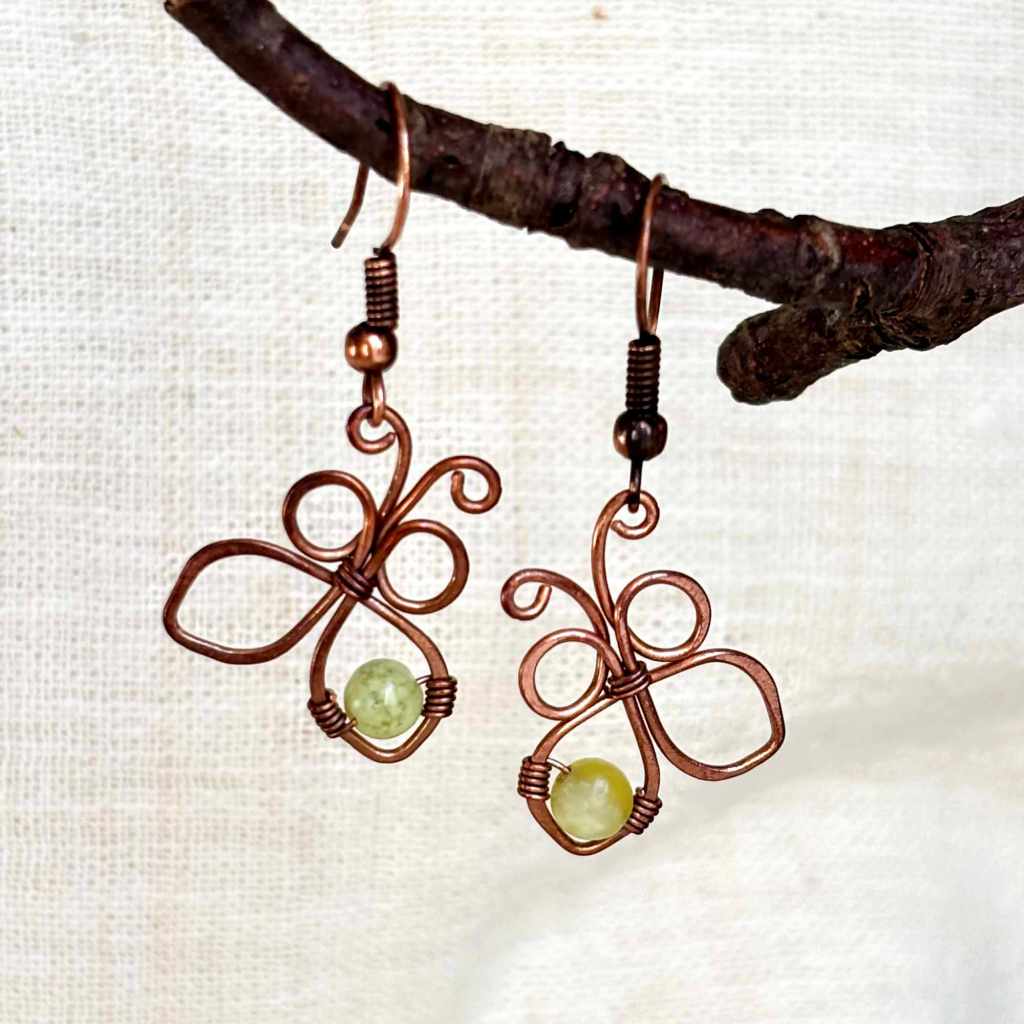 Handmade copper butterfly earrings hanging from a branch, showing the full wire wrapped design