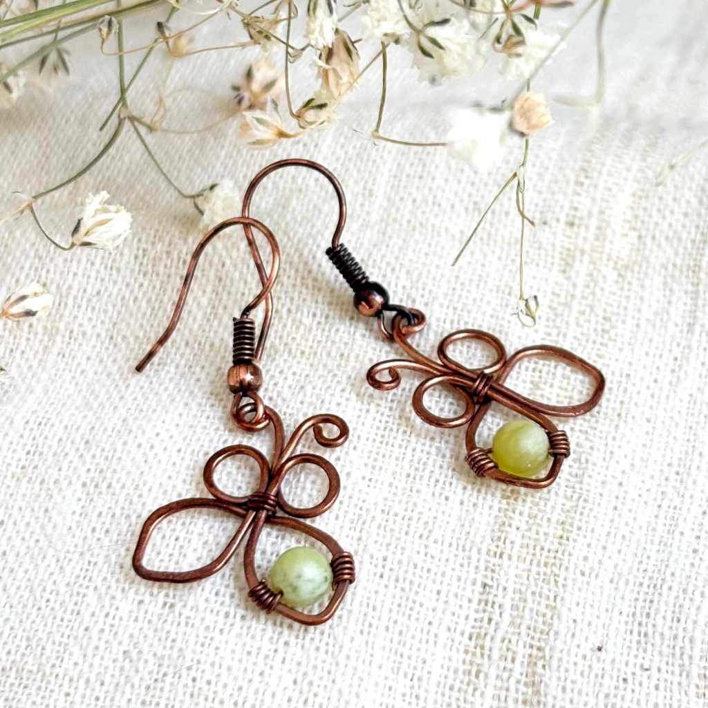 Handmade copper butterfly earrings with natural green stones, close-up view of wire wrapped design, shot on creme linen with dry flowers